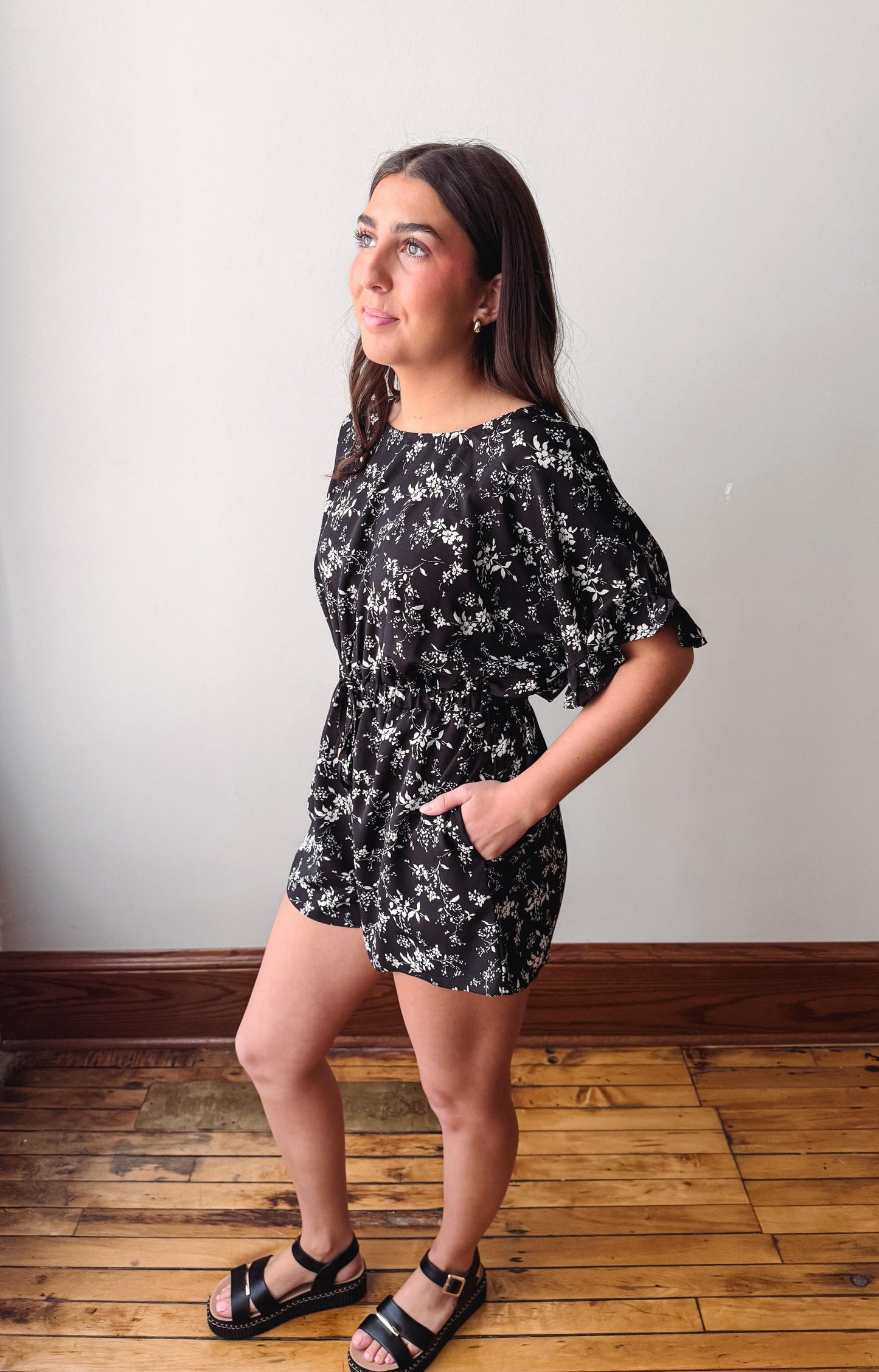 Woman wearing a black floral dress standing on a wooden floor against a white wall.