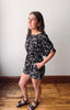 Woman wearing a black floral dress standing on a wooden floor against a white wall.