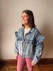 A woman wearing a light blue denim jacket with ruffle details on the shoulders and sleeves, paired with pink pants, standing in an indoor setting against a plain beige wall.