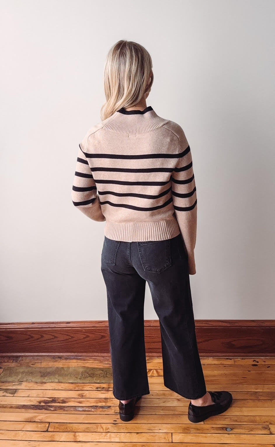 Person wearing a striped sweater and dark pants standing in a room with wooden flooring and a white wall.