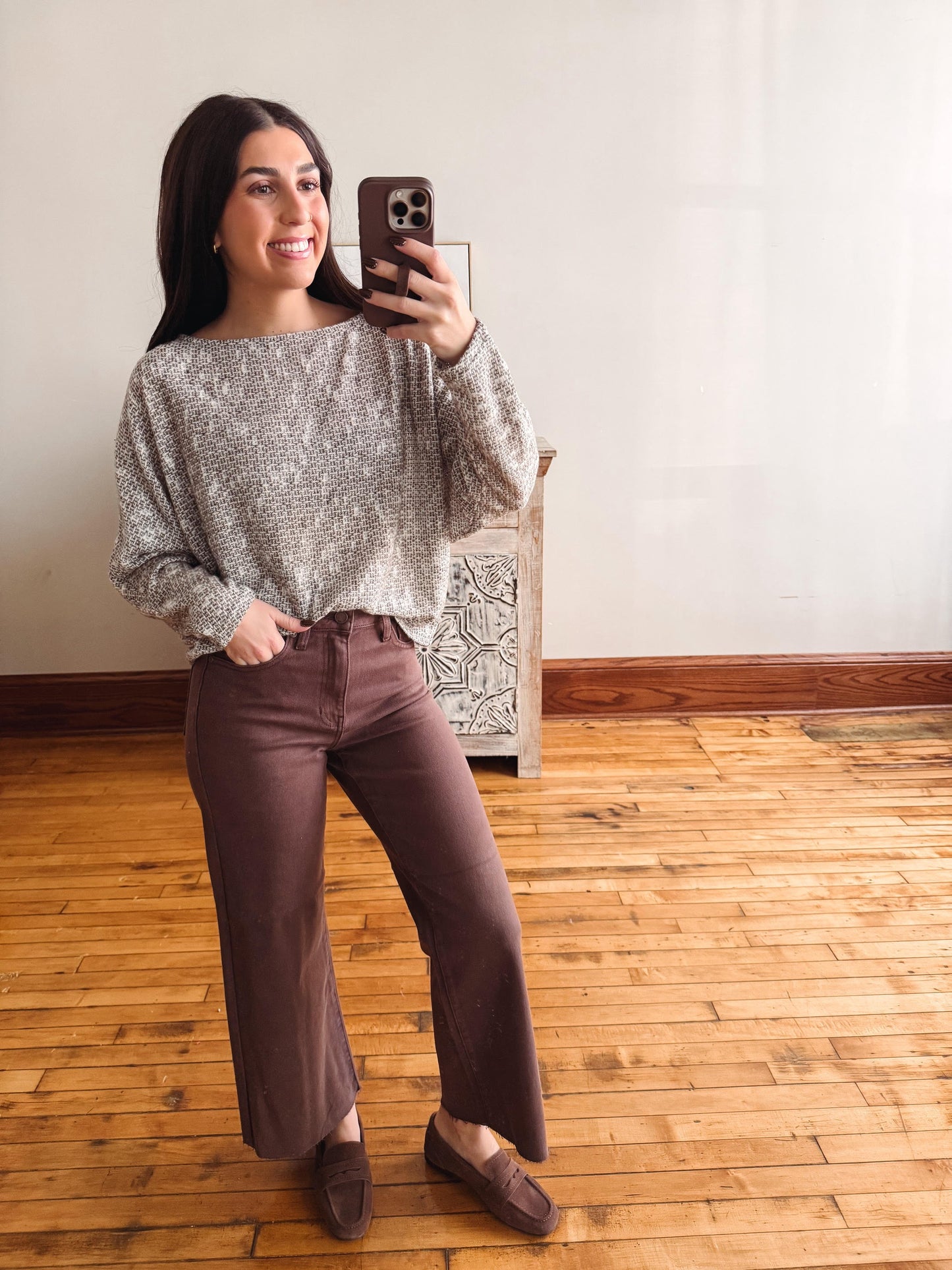 A woman takes a mirror selfie in a room with light wood floors and a white wall, wearing a textured gray knit top, brown wide-leg pants, and brown loafers.