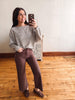 A woman takes a mirror selfie in a room with light wood floors and a white wall, wearing a textured gray knit top, brown wide-leg pants, and brown loafers.