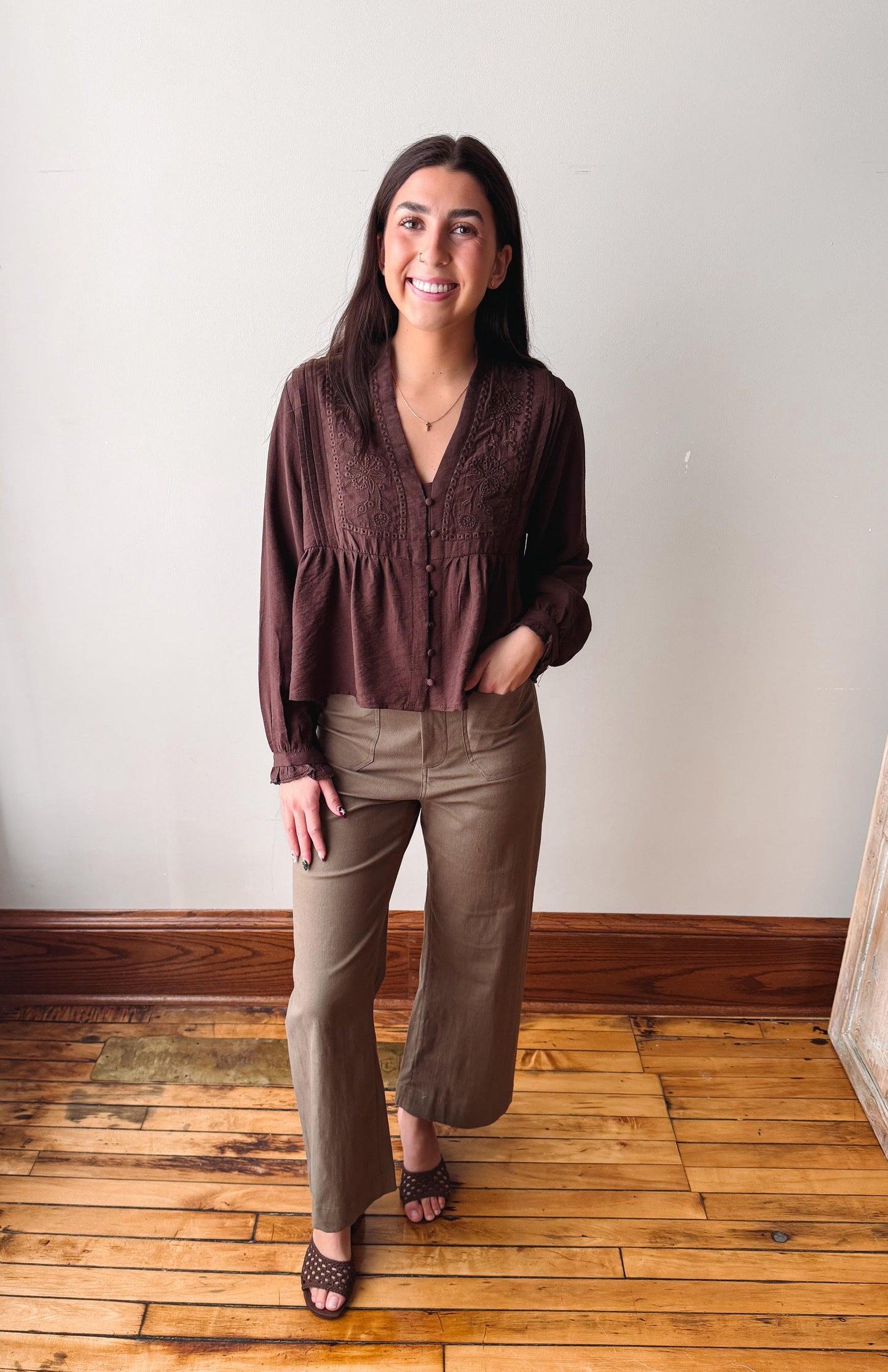 A woman modeling a dark brown long-sleeve lace-trim blouse and olive green wide-leg pants, standing on a wooden floor against a plain off-white wall.