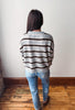 A woman viewed from behind wearing a gray and brown horizontally striped knit sweater, blue jeans, and brown shoes, standing on a wooden floor against a plain off-white wall.
