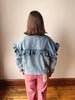 A person viewed from behind wearing a light blue denim jacket with dramatic ruffled shoulder details and pink pants, standing against a plain beige wall on a wooden floor.