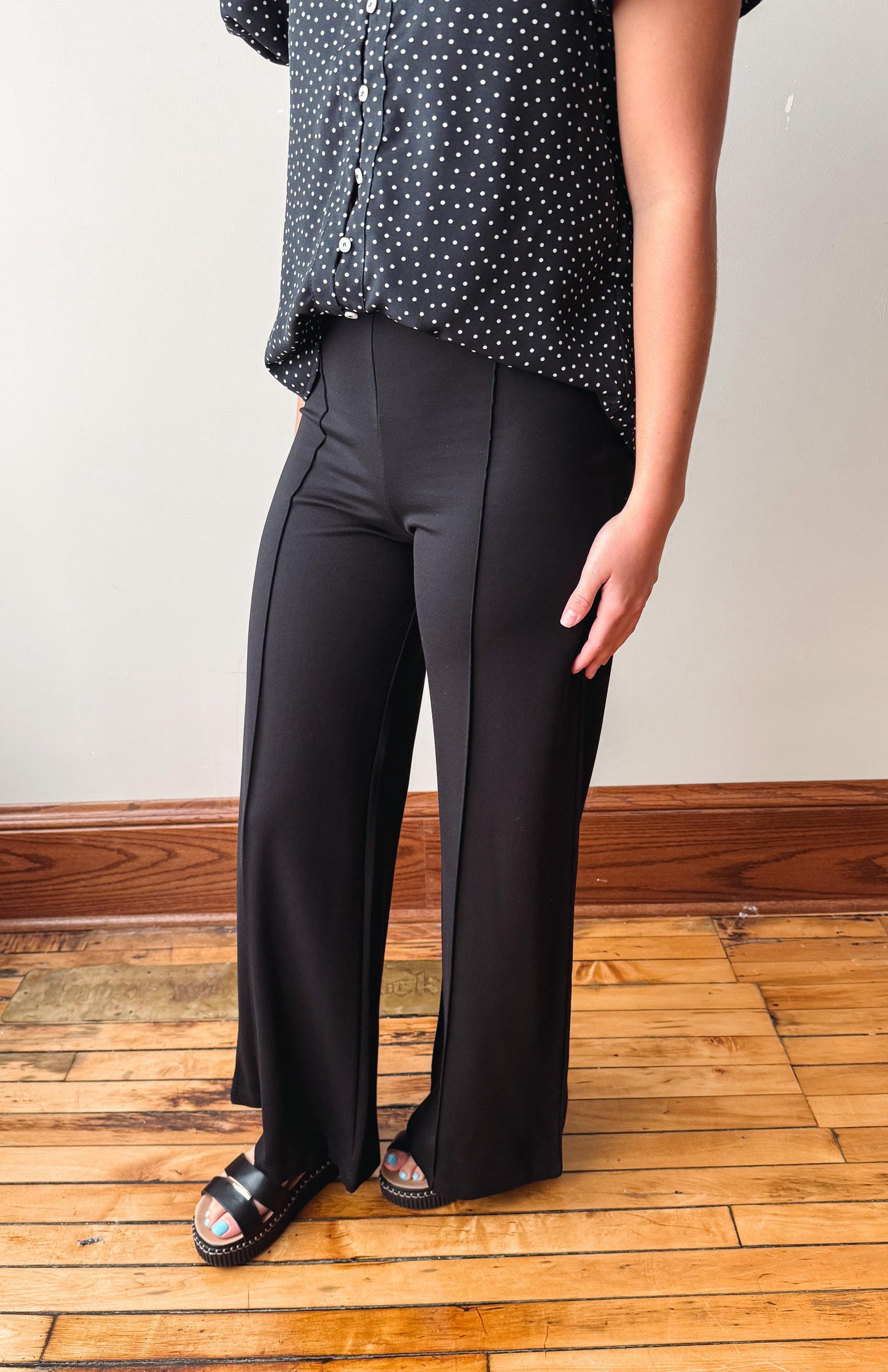A person wearing black wide-leg pants with a black and white polka dot blouse, standing on a wooden floor against a light wall.