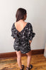 Woman wearing a black floral romper standing on a wooden floor with a white wall background
