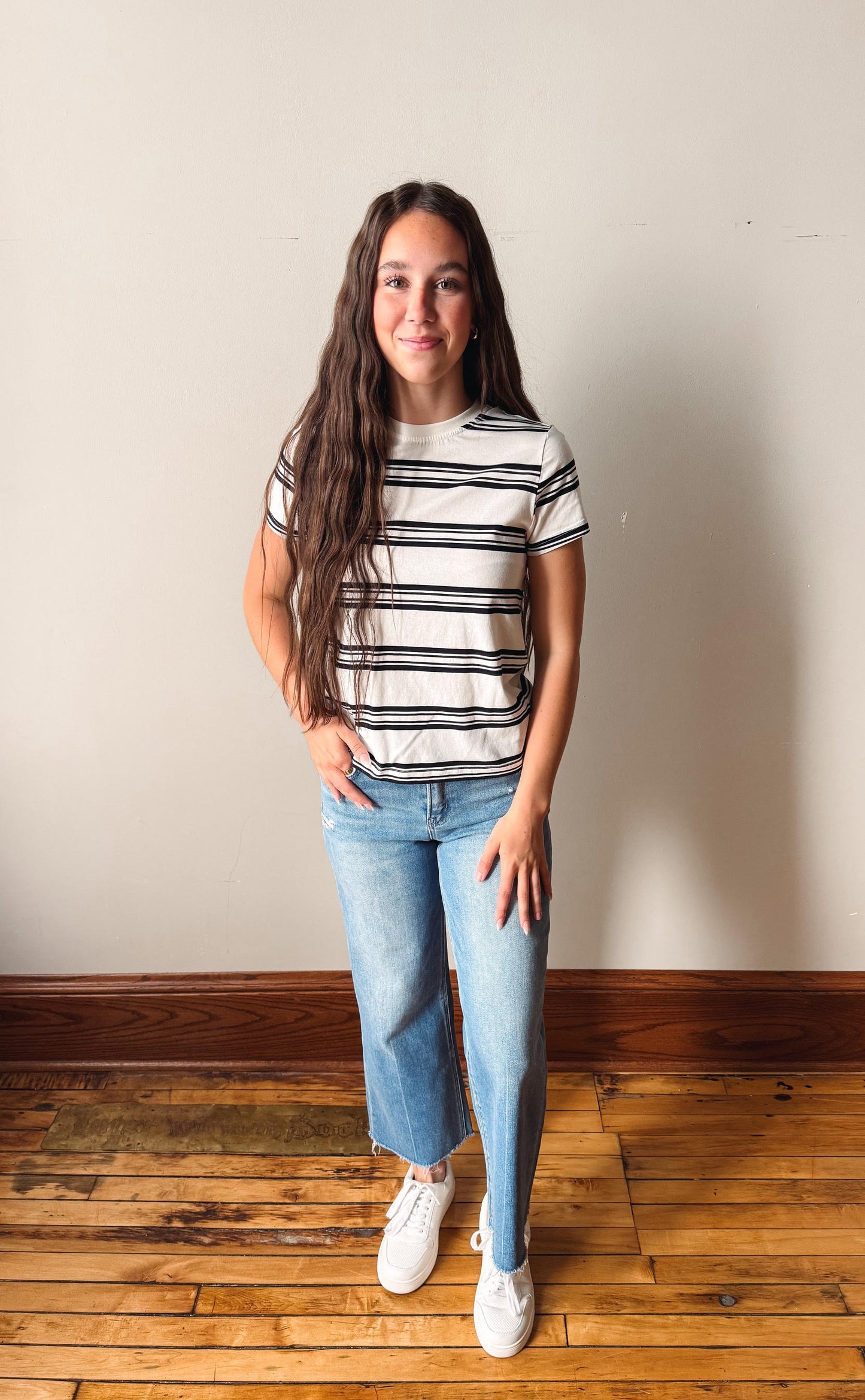 Woman wearing a striped shirt and jeans standing on a wooden floor with a plain wall background