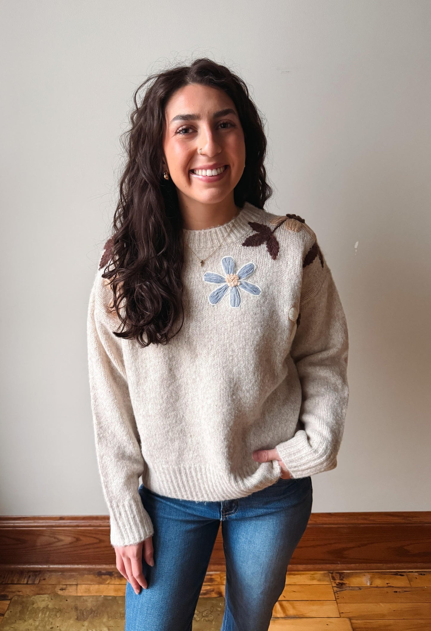A smiling woman with long dark wavy hair stands indoors wearing a cream knit sweater with a blue flower and brown leaf motif on the chest, paired with blue jeans.