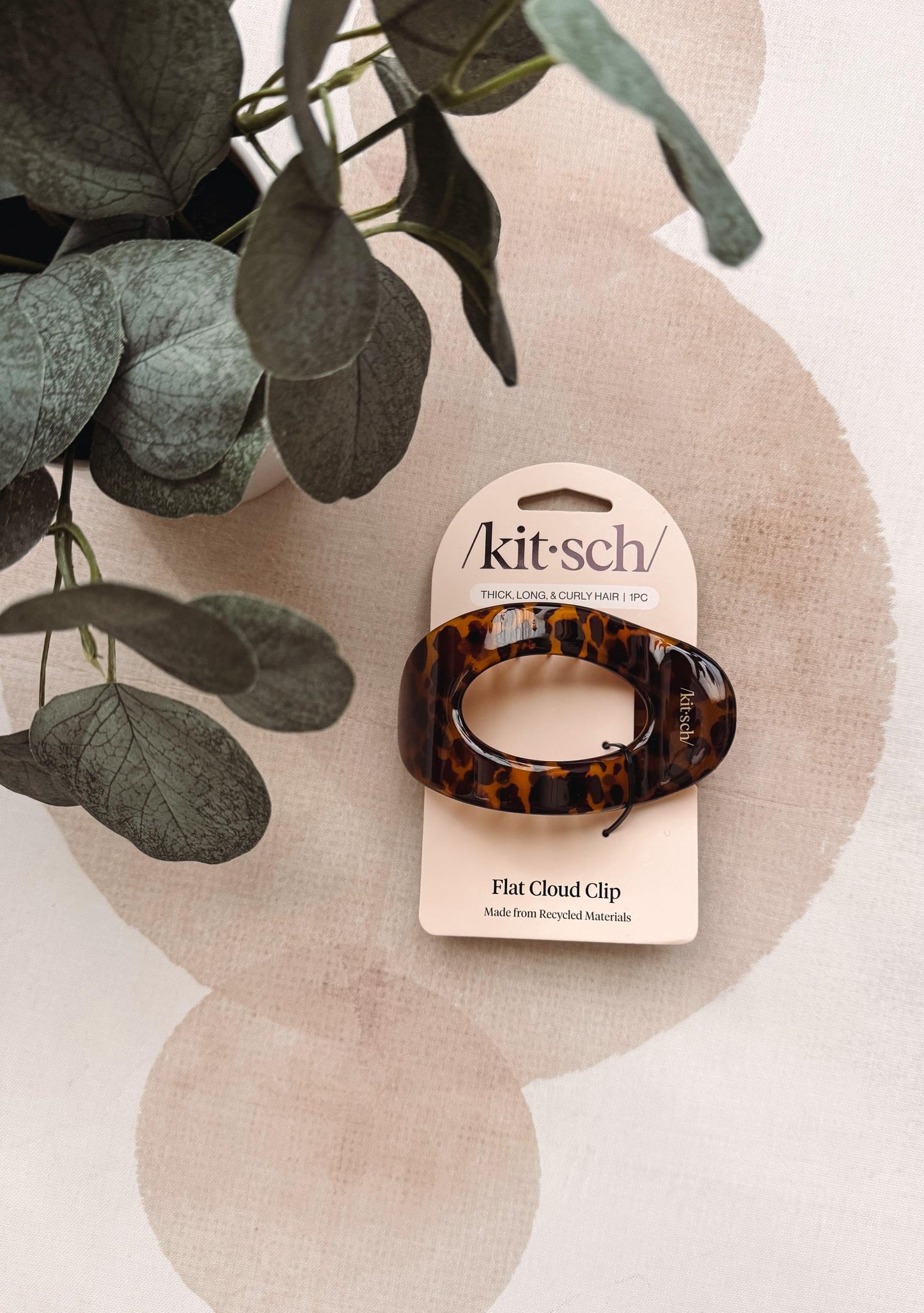 A tortoiseshell flat cloud hair clip by kitsch on a beige card, styled with green eucalyptus leaves on a patterned beige background.