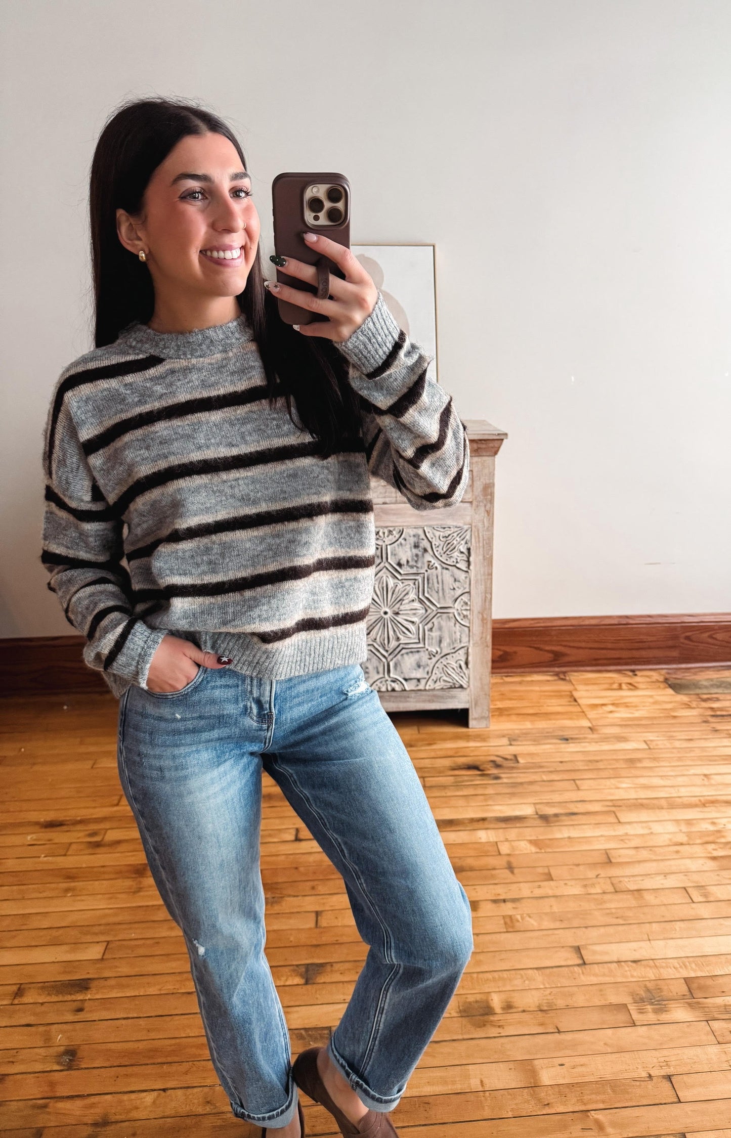 A woman takes a mirror selfie wearing a gray and black horizontally striped knit sweater, blue jeans, and brown shoes in a room with light-colored walls and wooden floors.