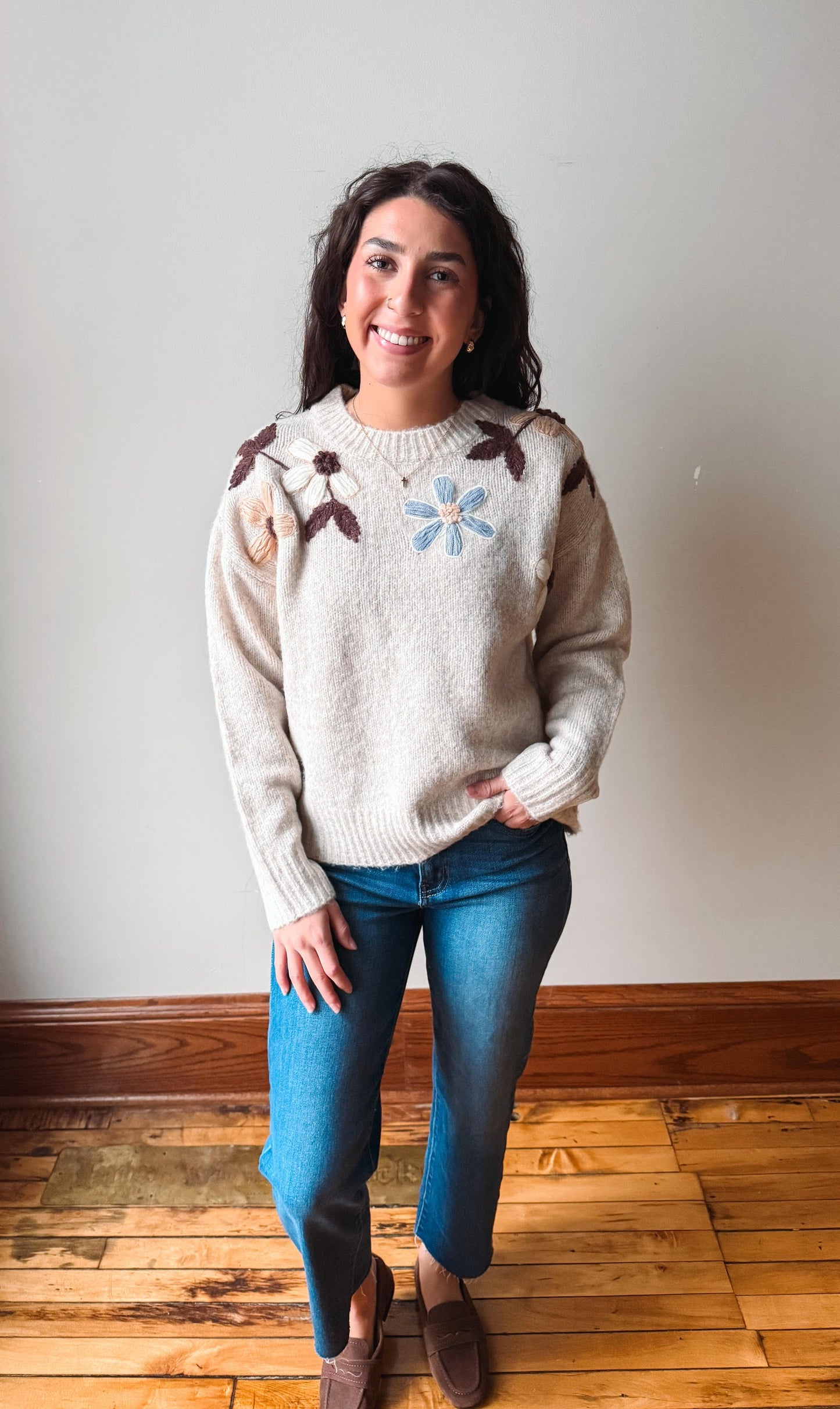 A woman wearing a cream knit sweater with brown leaf details and a light blue flower on the chest, paired with blue jeans and brown slip-on shoes, standing in a room with a light gray wall and wooden floor.
