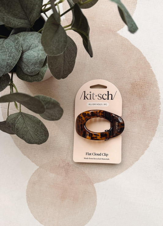 A tortoiseshell flat cloud clip by kitsch on its original cream-colored card packaging, styled on a textured beige surface with a green eucalyptus plant in the background.