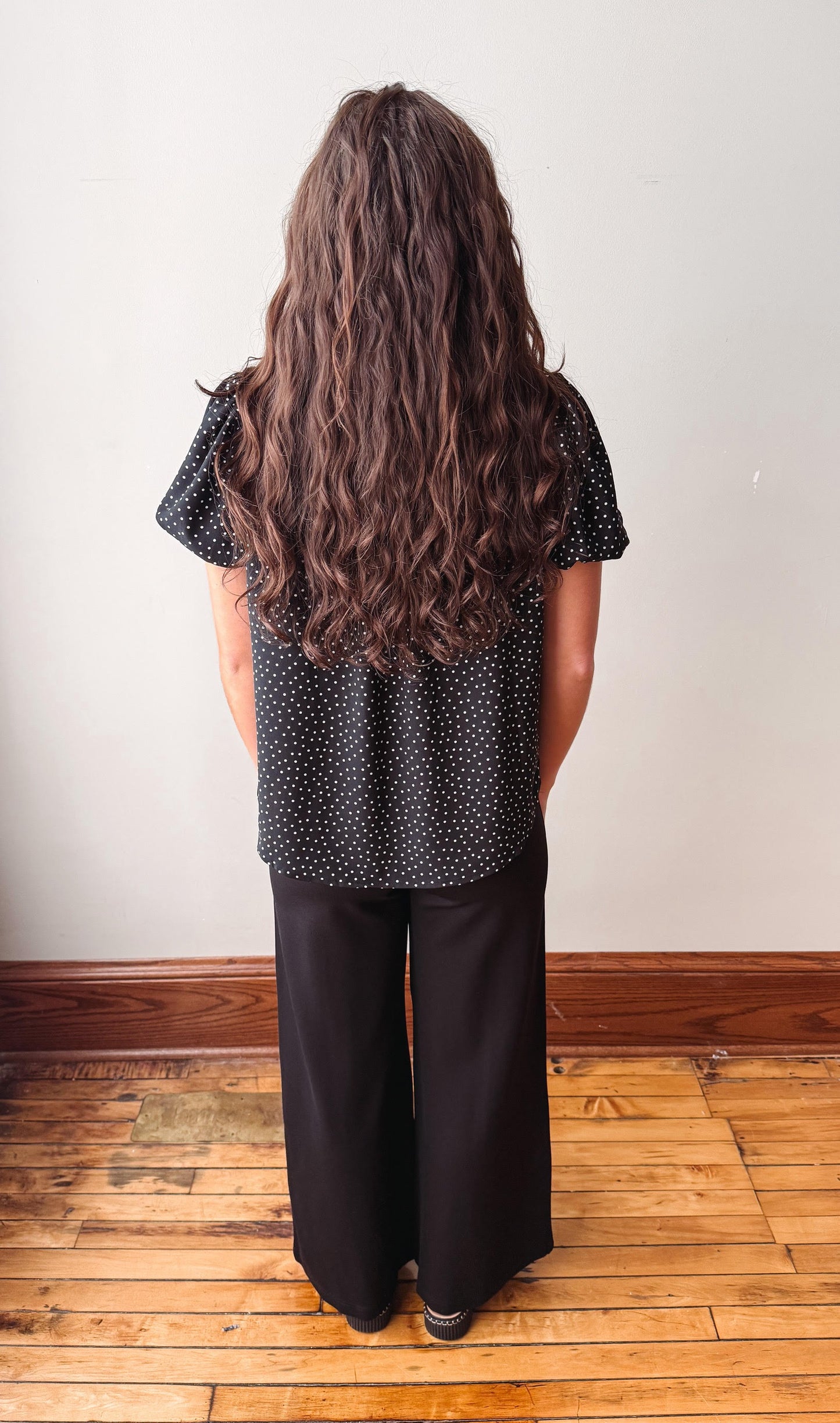 A woman with long, wavy brown hair is shown from behind, wearing a black short-sleeve top with white polka dots and black wide-leg pants, standing on a wooden floor against a plain off-white wall.