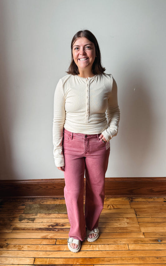 A woman standing in a room with a light gray wall and wooden floor, wearing a cream ribbed long-sleeve henley top and dusty rose pants.