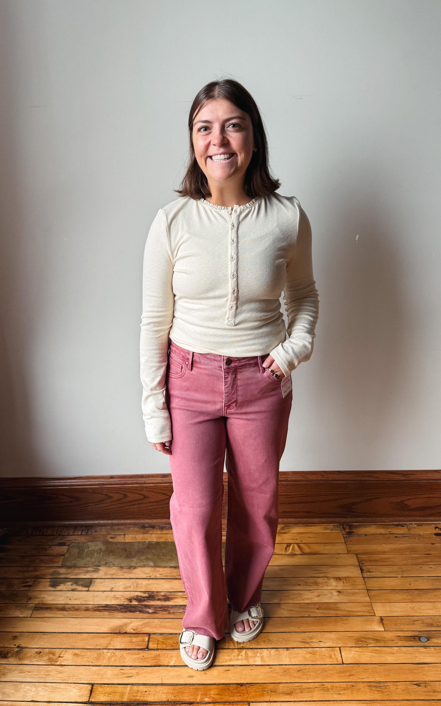 A woman standing in a room with a light gray wall and wooden floor, wearing a cream ribbed long-sleeve henley top and dusty rose pants.