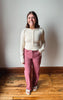 A woman standing in a room with a light gray wall and wooden floor, wearing a cream ribbed long-sleeve henley top and dusty rose pants.