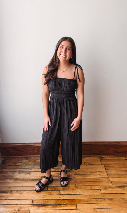 Woman wearing a black jumpsuit standing on a wooden floor with a white wall background