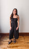 Woman wearing a black jumpsuit standing on a wooden floor with a white wall background