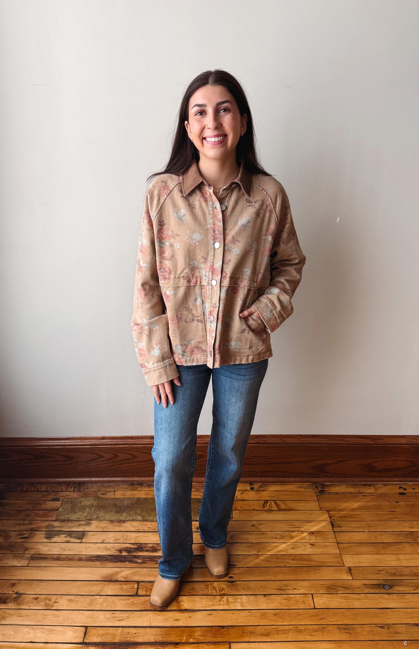 A woman stands in a room with a light gray wall and wooden floor, wearing a beige button-up shirt with a subtle floral pattern, blue jeans, and tan boots.