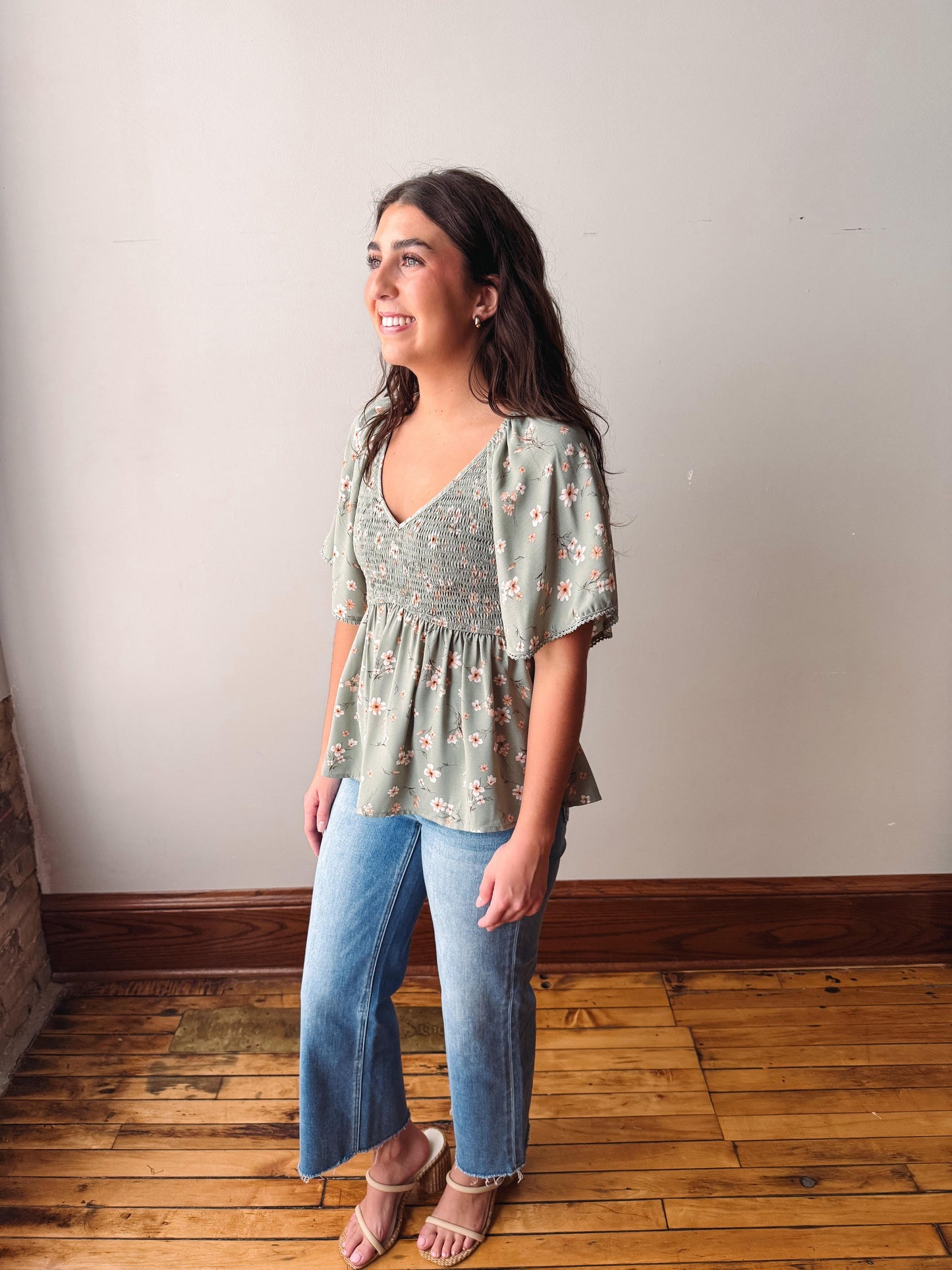 Woman wearing a green floral blouse and blue jeans standing against a plain wall.