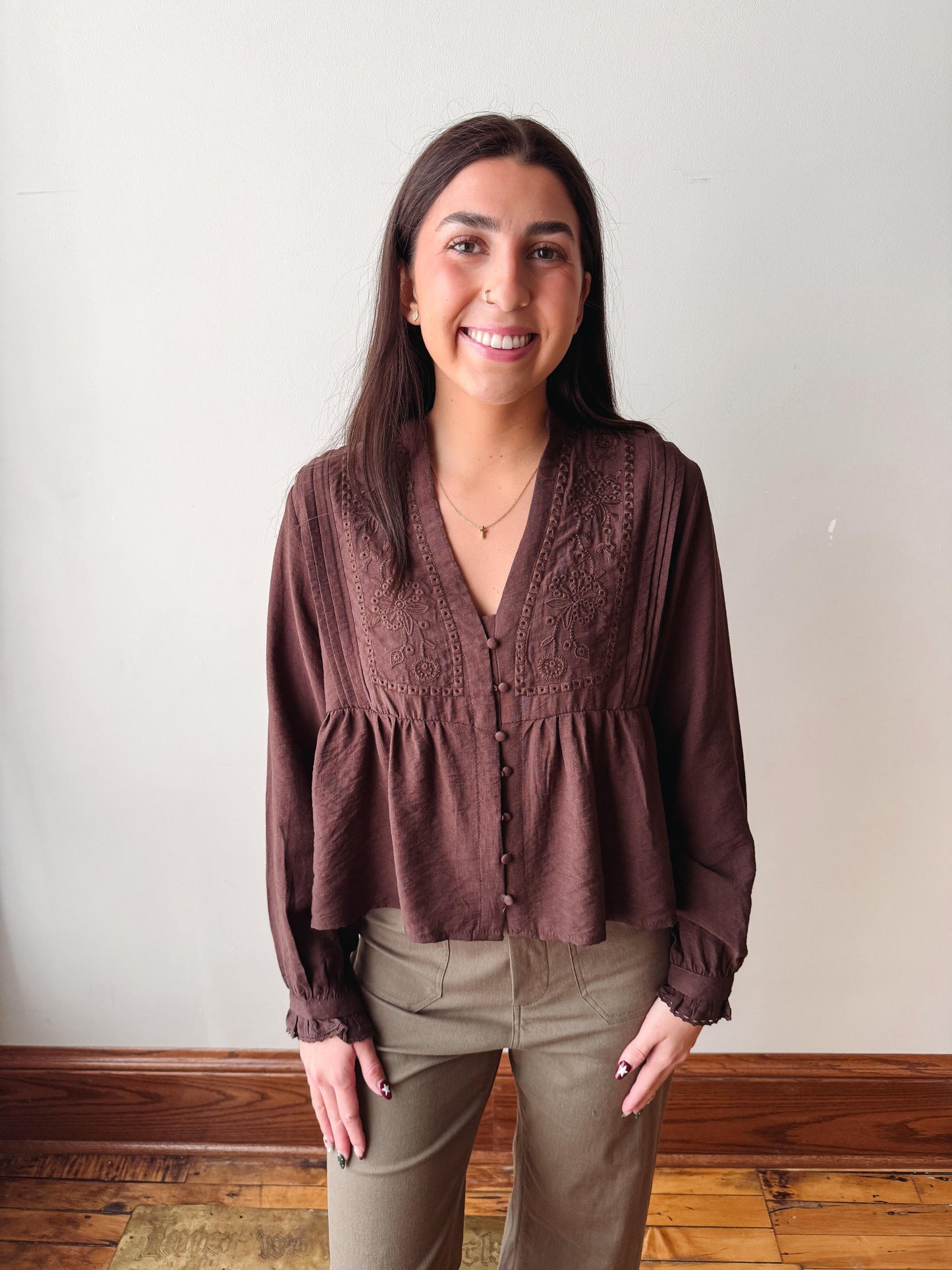 A woman smiling and wearing a brown long-sleeve button-front blouse with lace detailing on the chest and gathered cuffs, paired with olive green pants, standing against a plain off-white wall.