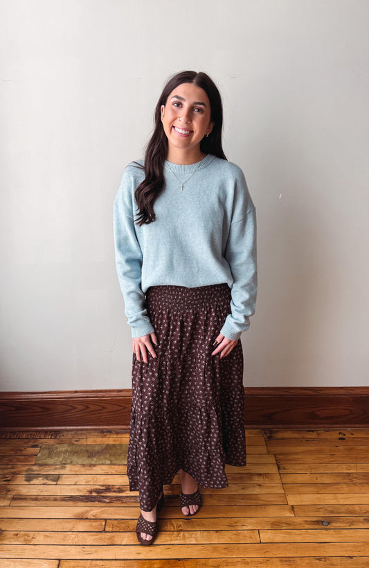A woman stands indoors against a plain light wall on a wooden floor, wearing a light blue long-sleeve knit sweater and a dark brown midi skirt with a small white floral pattern.