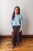 A woman stands indoors against a plain light wall on a wooden floor, wearing a light blue long-sleeve knit sweater and a dark brown midi skirt with a small white floral pattern.