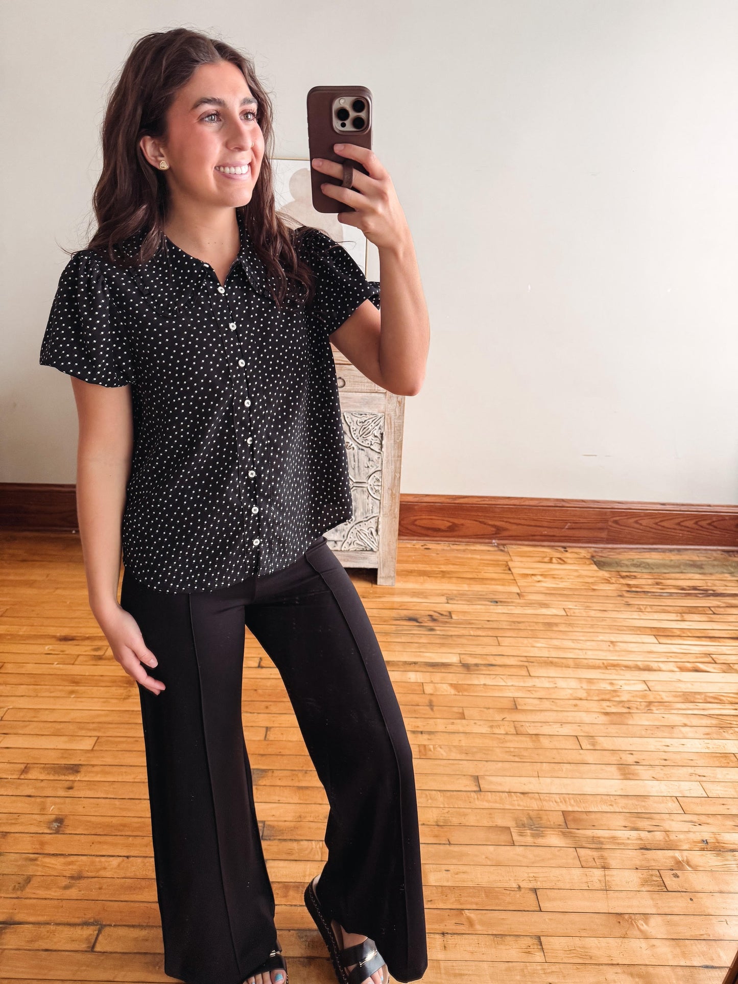 A woman taking a mirror selfie in a room with hardwood floors and a white wall, wearing a black short-sleeve button-up blouse with a white polka dot pattern and black wide-leg pants.