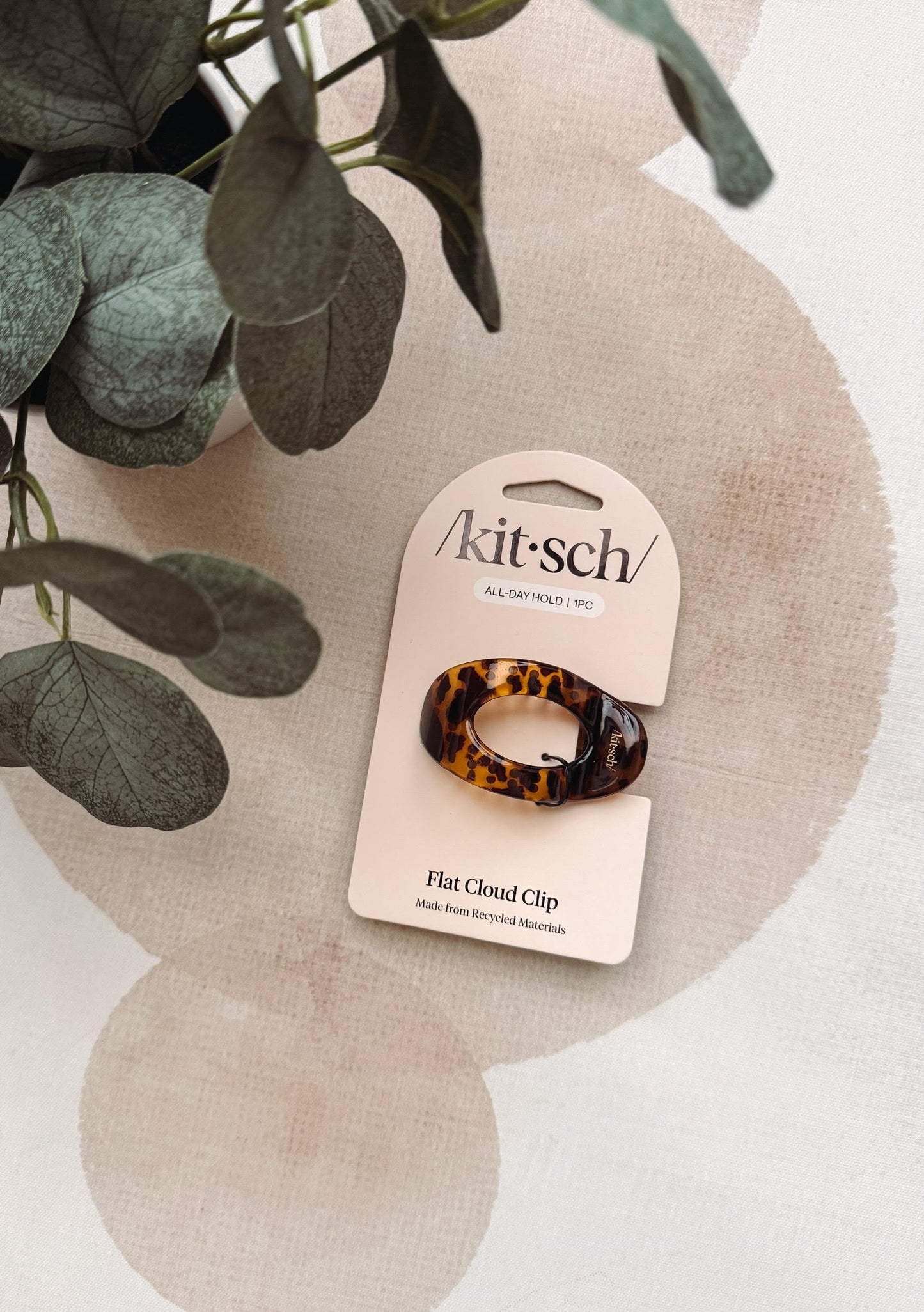A tortoiseshell flat cloud clip in its original packaging card, styled on a textured beige surface with a plant leaf in the corner.