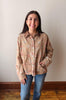 A woman smiling while wearing a beige button-up shirt with a pink and blue floral pattern, paired with blue jeans, standing against a plain beige wall with a wooden baseboard and floor.