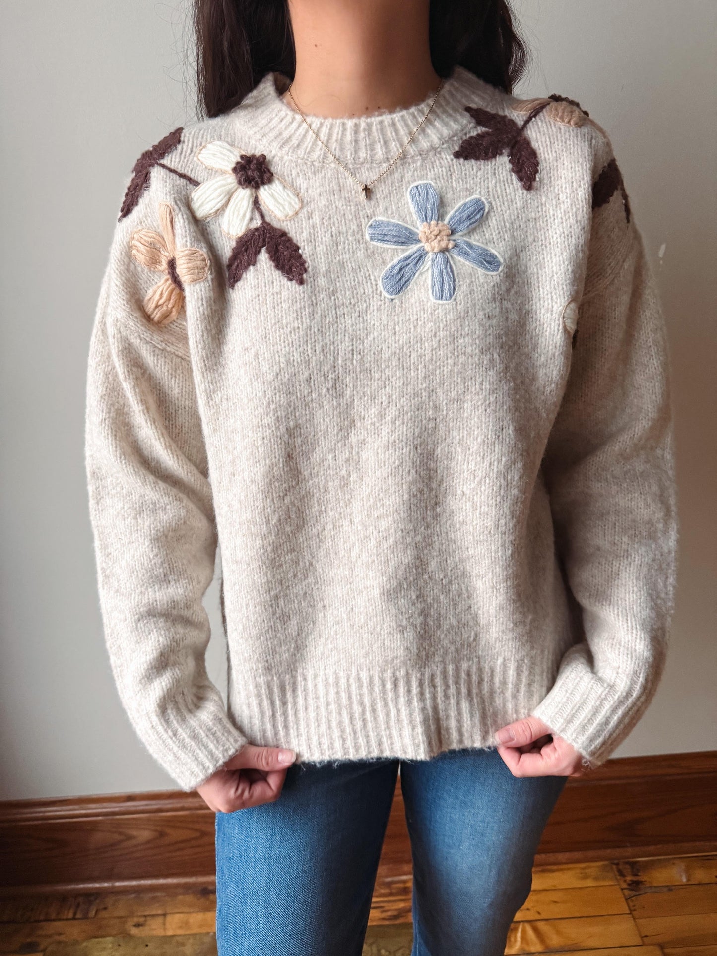 A person wearing a cream-colored knit sweater with raised floral embroidery on the chest and shoulders, paired with blue jeans, standing indoors against a plain wall.