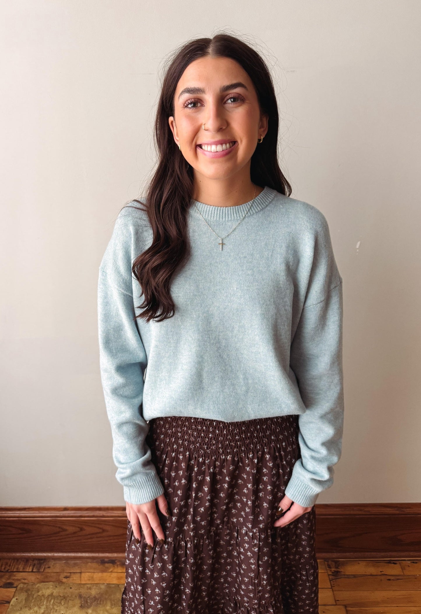 A woman with long dark hair stands indoors wearing a light blue long-sleeve knit sweater and a brown patterned skirt against a plain beige wall.