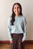A woman with long dark hair stands indoors wearing a light blue long-sleeve knit sweater and a brown patterned skirt against a plain beige wall.
