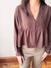 A woman wearing a brown long-sleeve blouse with intricate embroidery and button details, paired with khaki pants, standing indoors against a light wall.
