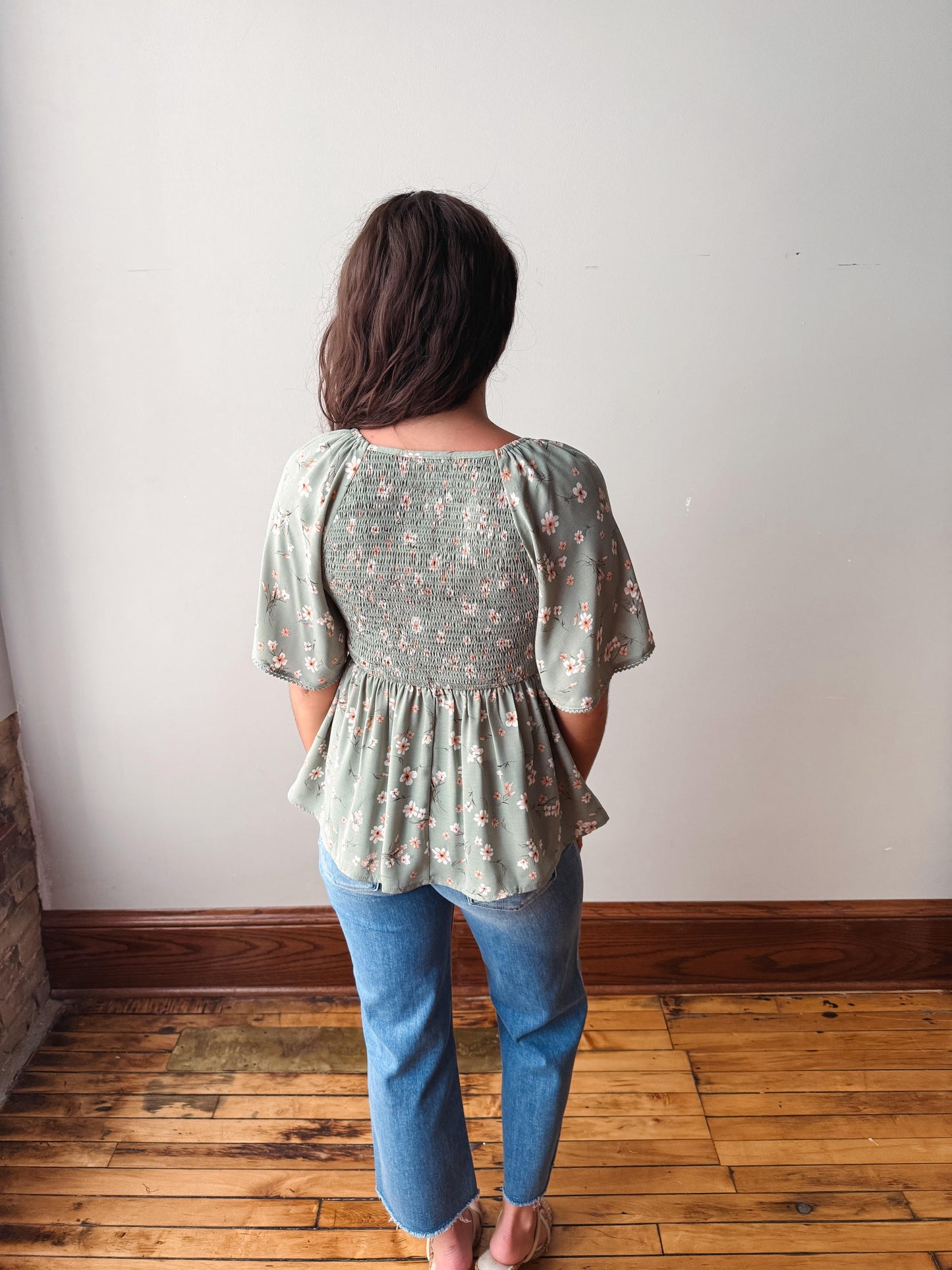 Person wearing a floral blouse and blue jeans standing against a white wall.