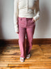 A person wearing a cream ribbed long-sleeve henley top and dusty rose pink jeans, standing on a wooden floor against a plain wall.