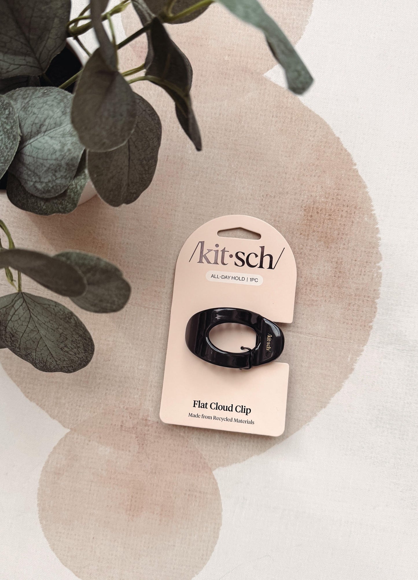 A black flat cloud clip by Kit's on its original beige cardboard packaging, styled on a textured beige surface with a plant in the background.