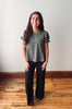 A woman stands in a room with a light gray wall and wooden floor, wearing a sage green short-sleeve blouse with white polka dots and black wide-leg pants.