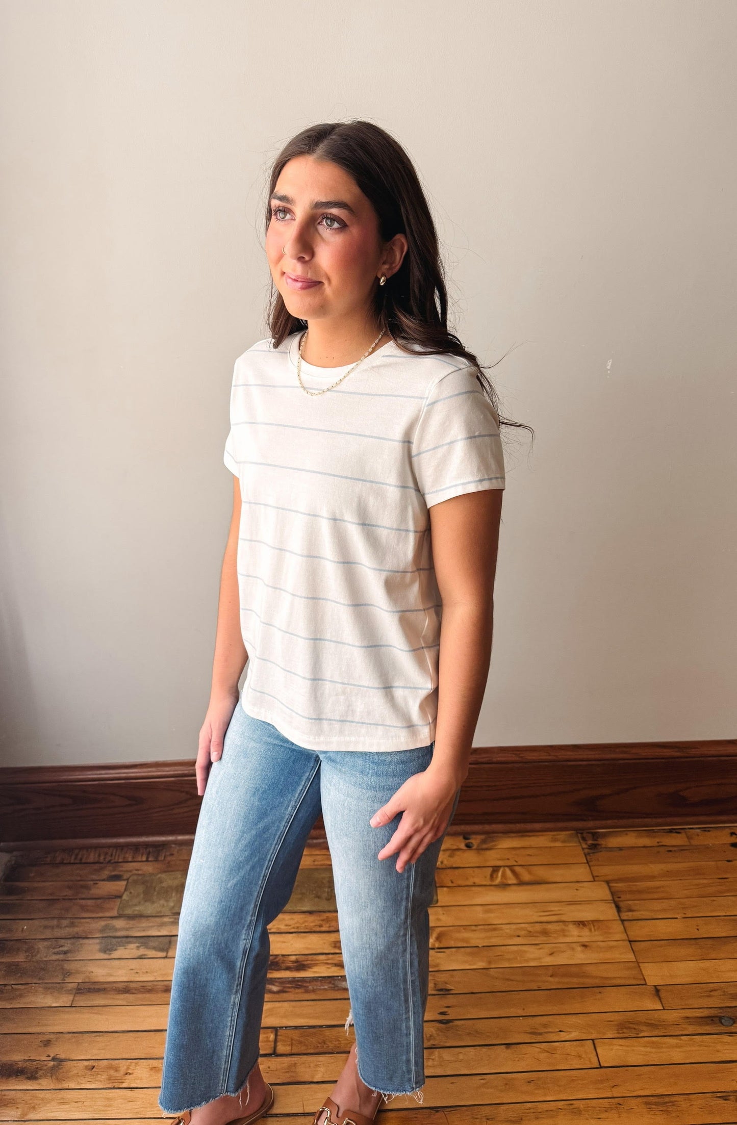 Woman wearing a striped shirt and jeans standing against a plain wall.