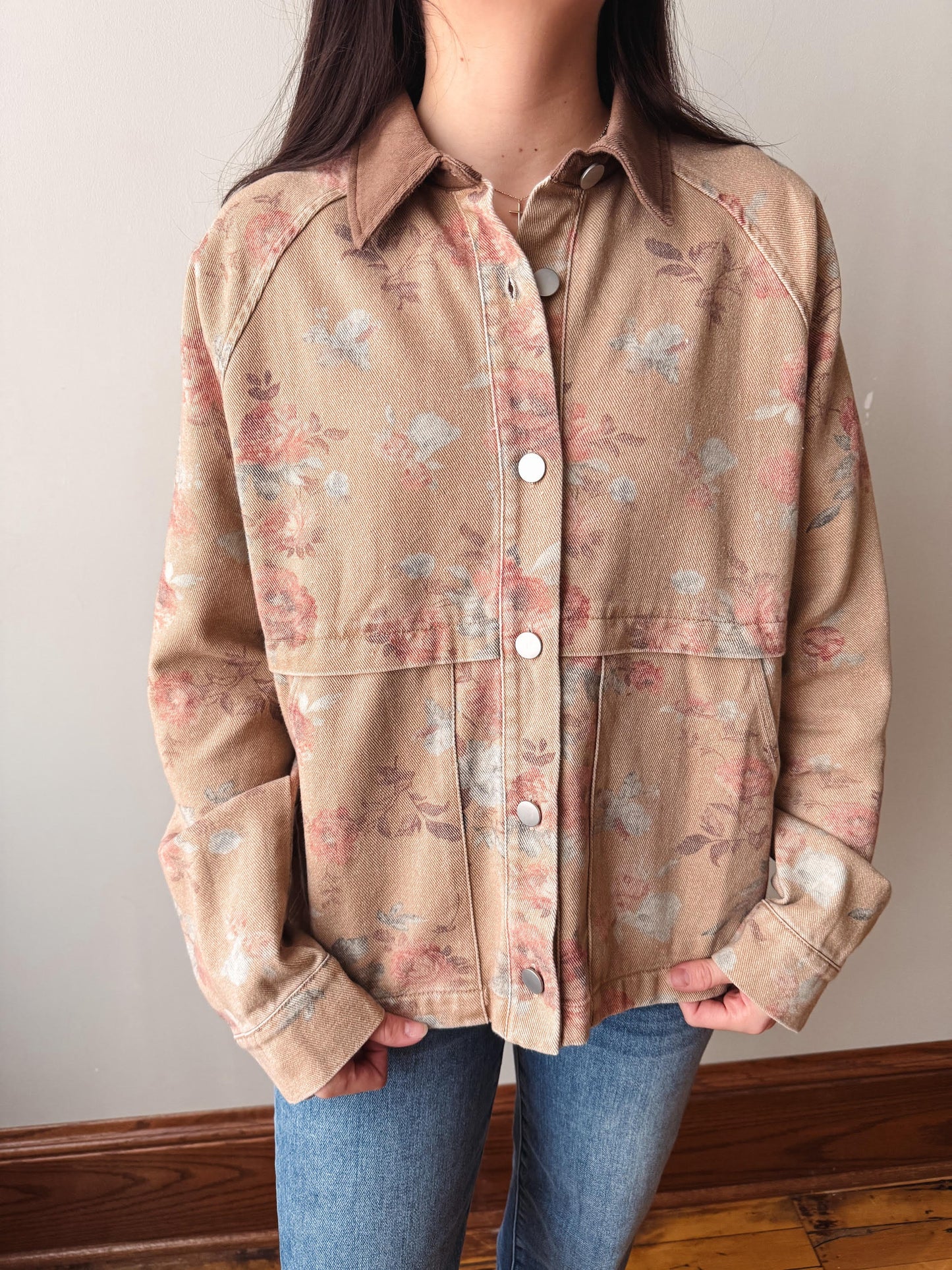 A person wearing a beige button-front shirt jacket with a muted pink and blue floral print, paired with blue jeans, standing indoors against a plain wall.