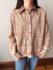 A person wearing a beige button-front shirt jacket with a muted pink and blue floral print, paired with blue jeans, standing indoors against a plain wall.
