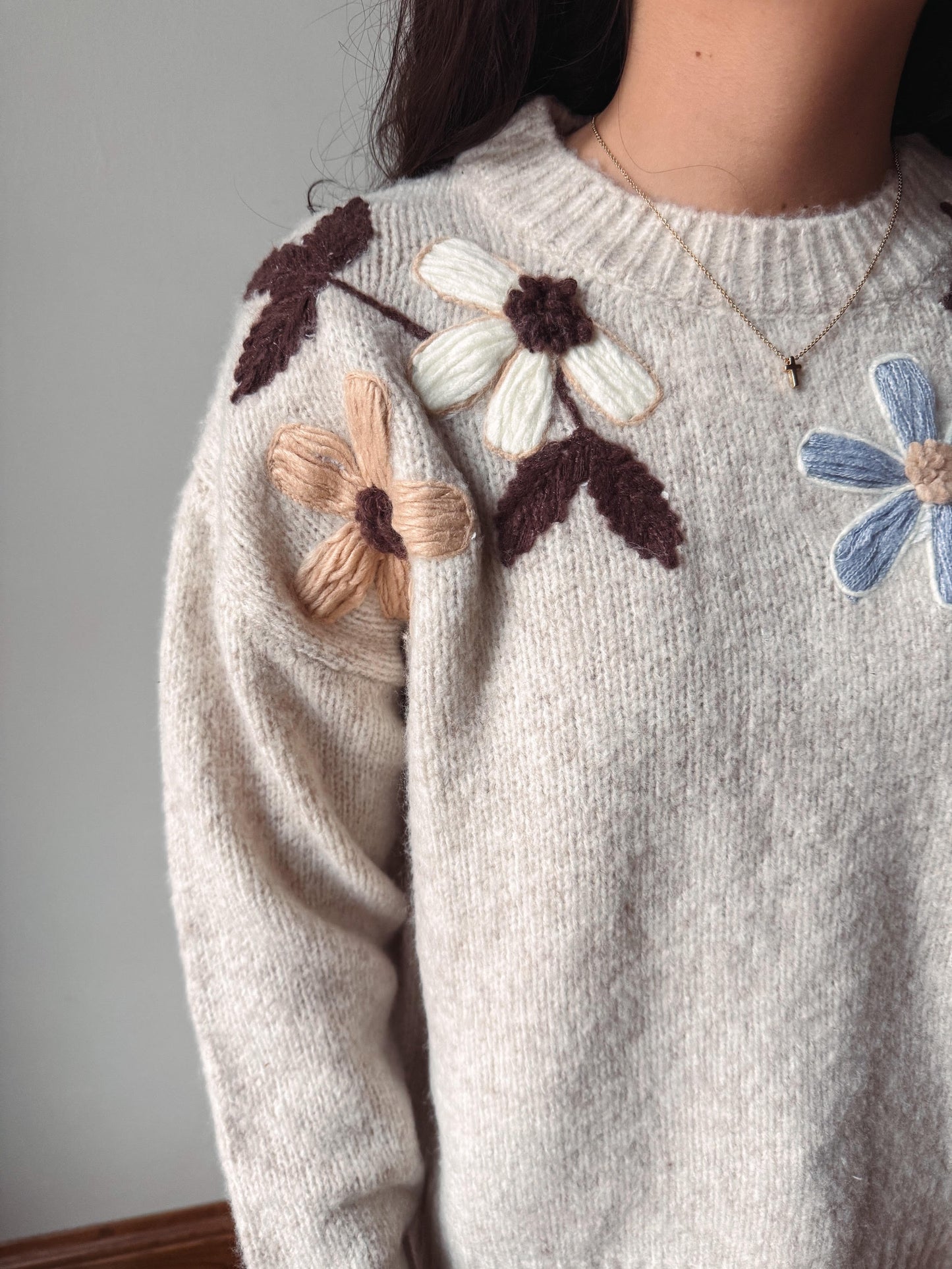 Close-up of a person wearing a cream knit sweater with large embroidered flowers in white, beige, and blue with dark brown leaves, paired with a delicate gold necklace.
