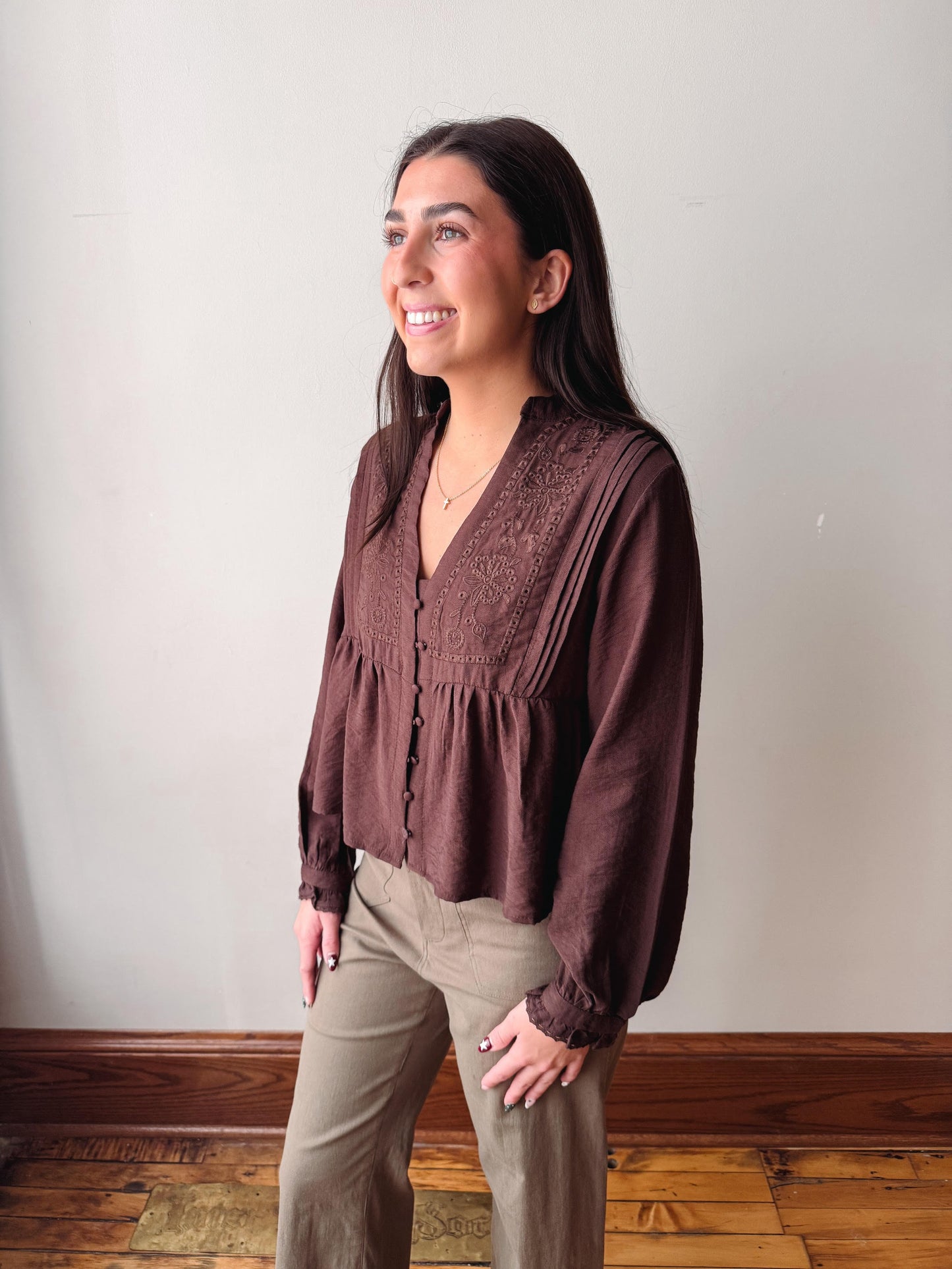 A woman modeling a brown long-sleeve blouse with a lace-trimmed V-neck and gathered peplum waist, paired with khaki pants, standing against a plain off-white wall.