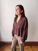 A woman modeling a brown long-sleeve blouse with a lace-trimmed V-neck and gathered peplum waist, paired with khaki pants, standing against a plain off-white wall.