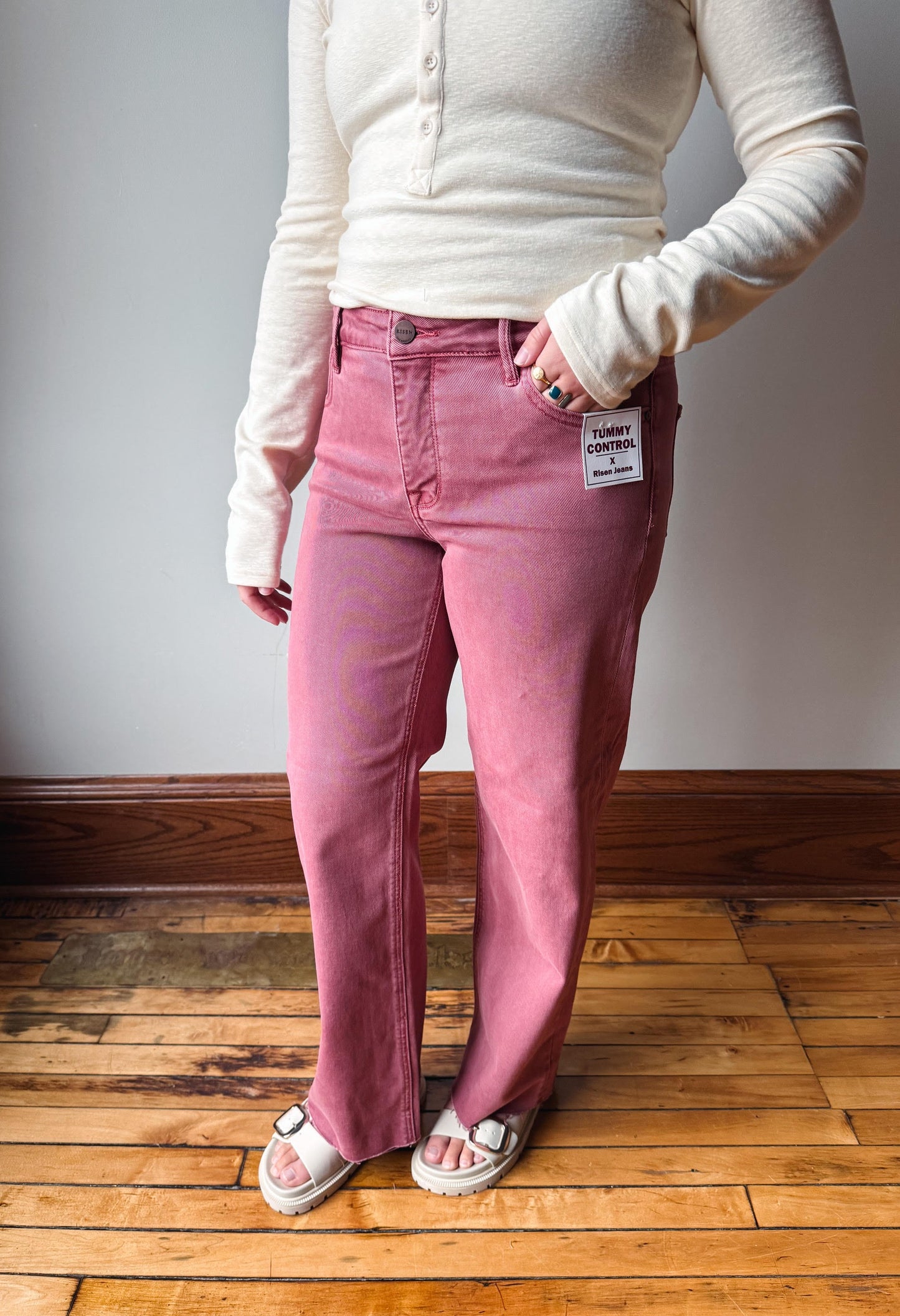 A person wearing a pair of pink denim jeans with a cream ribbed long-sleeve henley top and white slide sandals, standing on a wooden floor against a gray wall.