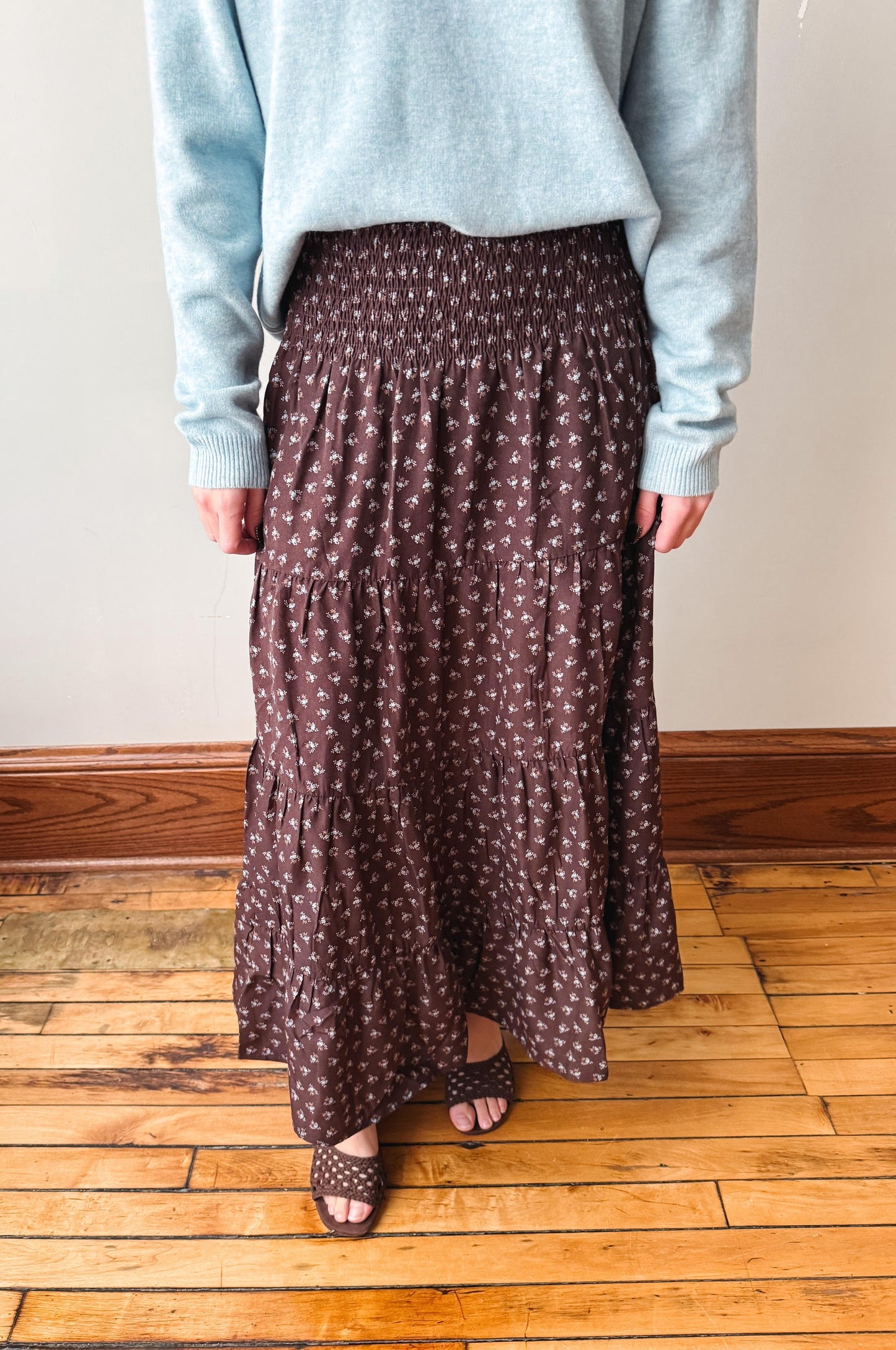 A person wearing a long brown tiered maxi skirt with a small white floral pattern, paired with a light blue sweater and brown sandals, standing on a wooden floor against a plain wall.
