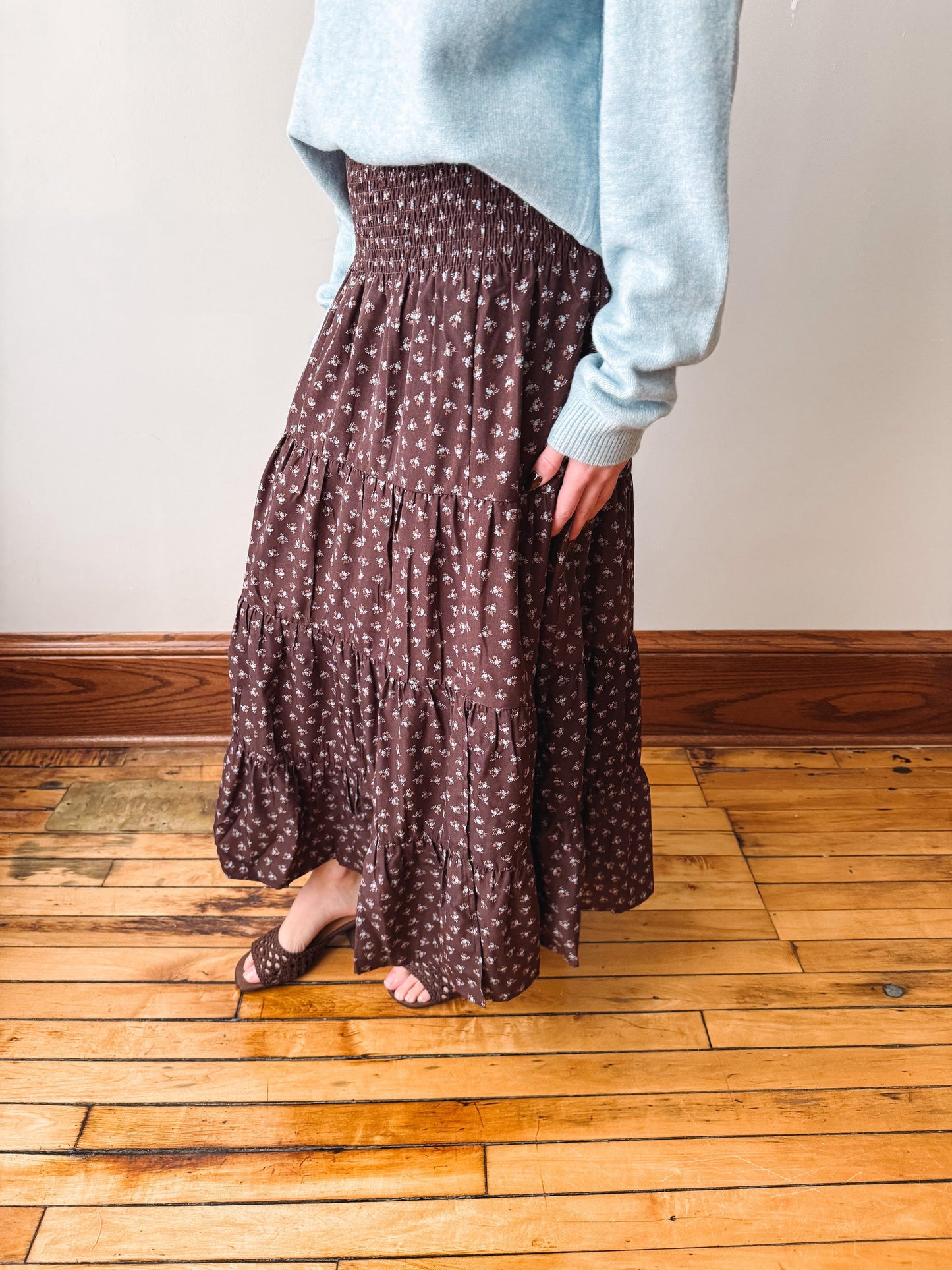 A person wearing a brown tiered maxi skirt with a small white floral pattern, paired with a light blue sweater and brown flat sandals, standing on a wooden floor against a beige wall.