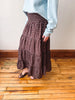 A person wearing a brown tiered maxi skirt with a small white floral pattern, paired with a light blue sweater and brown flat sandals, standing on a wooden floor against a beige wall.