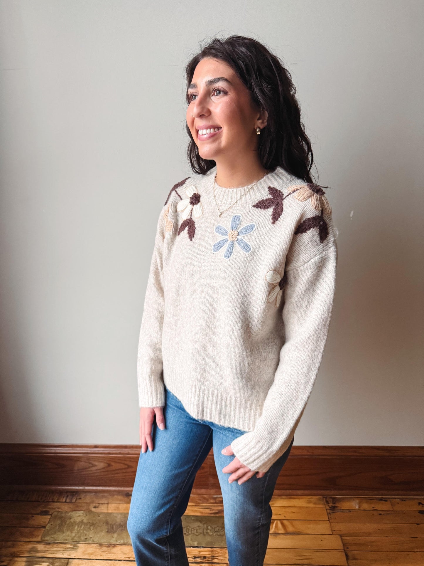 A woman wearing a cream-colored knit sweater with brown and light blue floral details on the shoulders and chest, paired with blue jeans, standing in a room with a neutral wall and wooden floor.